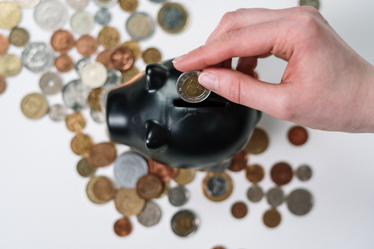 services-04 Close-up of a hand inserting a coin into a black piggy bank with scattered coins on a white background.
